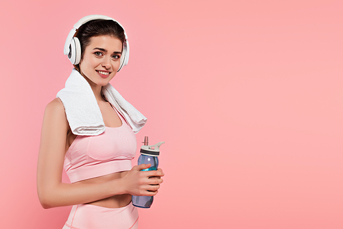 Sportswoman smiling at camera while listening music in headphones and holding sports bottle isolated on pink