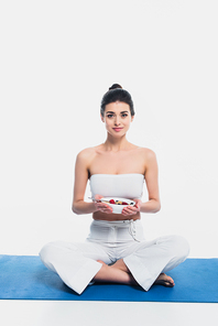Woman with crossed legs holding bowl of cereals with berries on fitness mat isolated on white