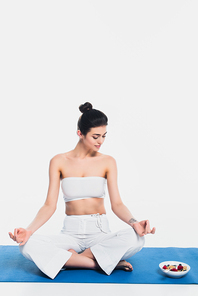 Young woman meditating near bowl of cereals with berries on fitness mat on white background
