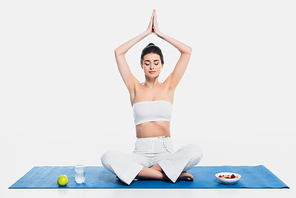 Brunette woman meditating near cereals, apple and water on fitness mat on white background