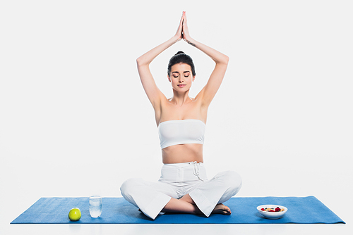 Brunette woman meditating near cereals, apple and water on fitness mat on white background