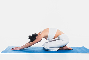 Side view of young woman sitting on fitness mat while practicing yoga isolated on white