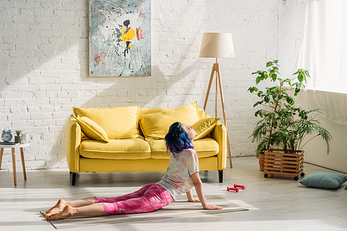 Girl with colorful hair in upward facing dog pose on yoga mat in living room