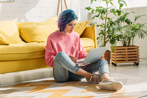 Freelancer with colorful hair working with laptop near sofa in living room