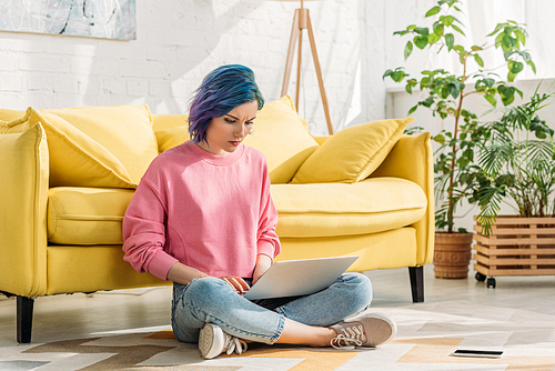 Freelancer with colorful hair working with laptop near smartphone on floor