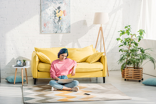 Freelancer with colorful hair and crossed legs working with laptop near sofa on floor