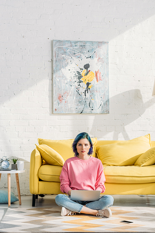 Freelancer with colorful hair and crossed legs  and working with laptop near sofa and smartphone on floor