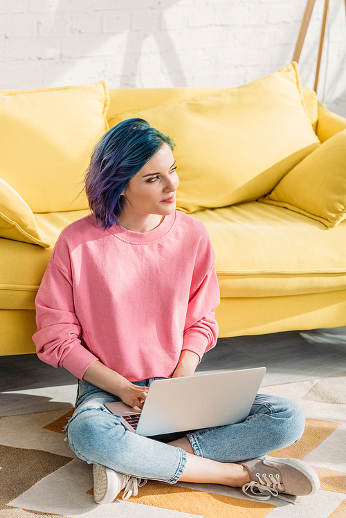 Thoughtful freelancer with colorful hair looking away and working with laptop near sofa on floor in living room