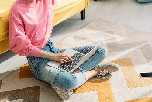 Cropped view of freelancer with crossed legs working with laptop near sofa and smartphone on floor in living room