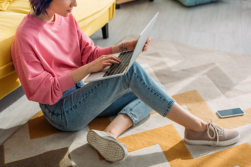 Cropped view of freelancer with colorful hair working with laptop near sofa and smartphone on floor in living room