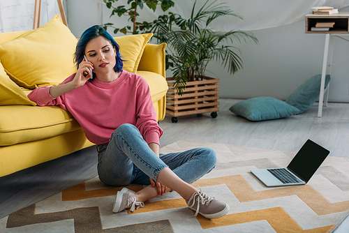 Freelancer with colorful hair and crossed legs talking on smartphone near sofa and laptop in living room