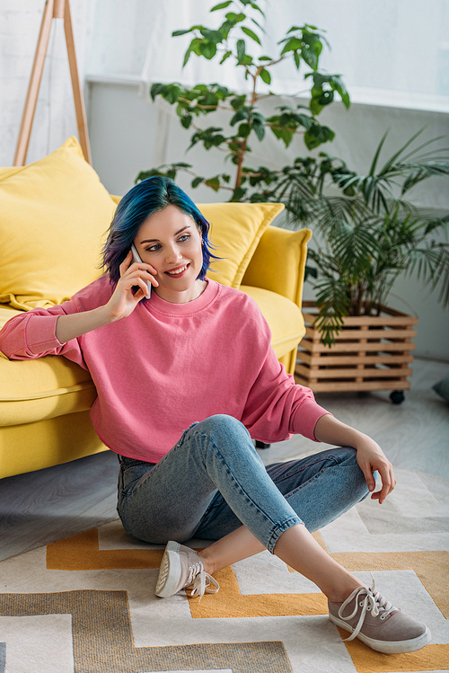 Woman with colorful hair and crossed legs smiling and talking on smartphone near sofa in living room