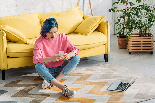 Freelancer with colorful hair and crossed legs holding smartphone on floor near sofa and laptop