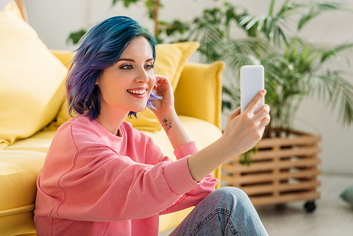 Girl with colorful hair smiling and taking selfie with smartphone near sofa on floor in living room