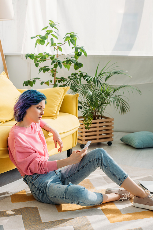 Concentrated woman with colorful hair holding smartphone near sofa on floor in living room