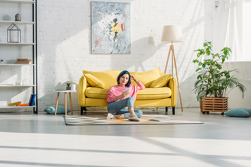 Woman with colorful hair and smartphone sitting near sofa on floor in living room