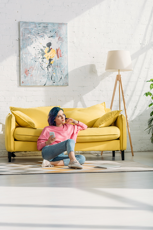 Thoughtful woman with colorful hair and smartphone sitting near sofa on floor in living room