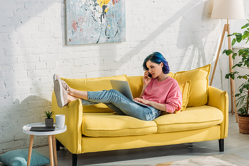 Freelancer with colorful hair talking on smartphone with laptop and lying on sofa