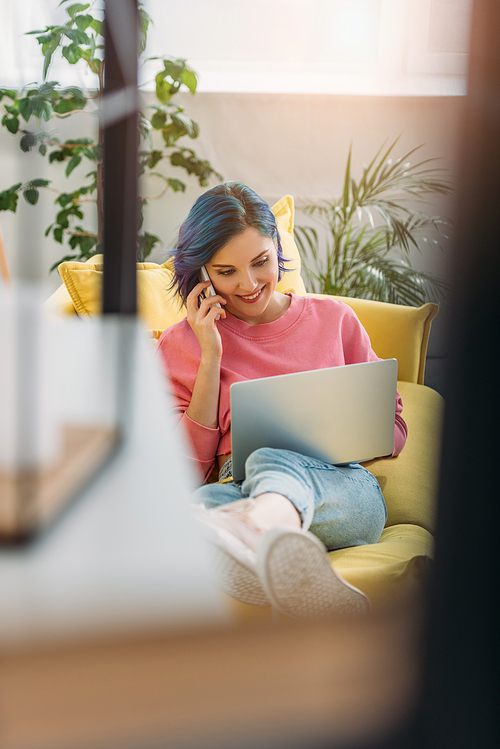 Selective focus of freelancer with colorful hair smiling and talking on smartphone with laptop on sofa