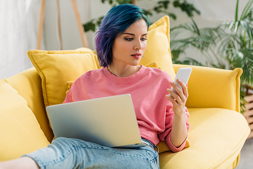 Freelancer with colorful hair and laptop looking at smartphone and lying on sofa