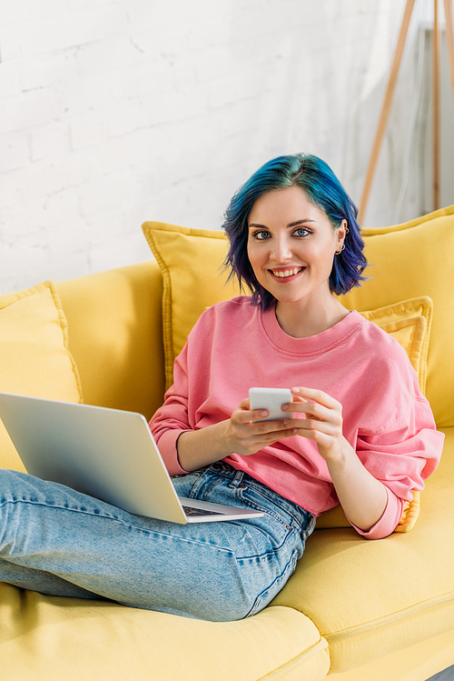 Freelancer with colorful hair, smartphone and laptop smiling and lying on sofa