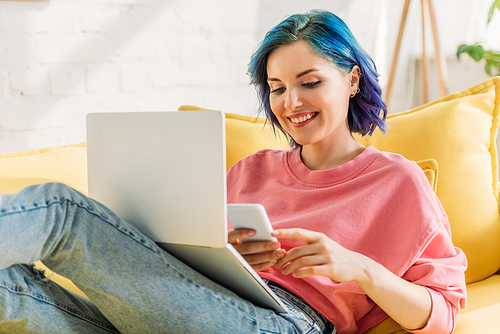 Freelancer with colorful hair and laptop holding smartphone, smiling and lying on sofa