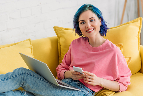 Freelancer with colorful hair and laptop holding smartphone, smiling,  and lying on sofa