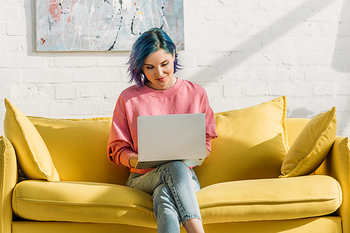 Freelancer with colorful hair and laptop smiling and sitting on yellow sofa with crossed legs in living room