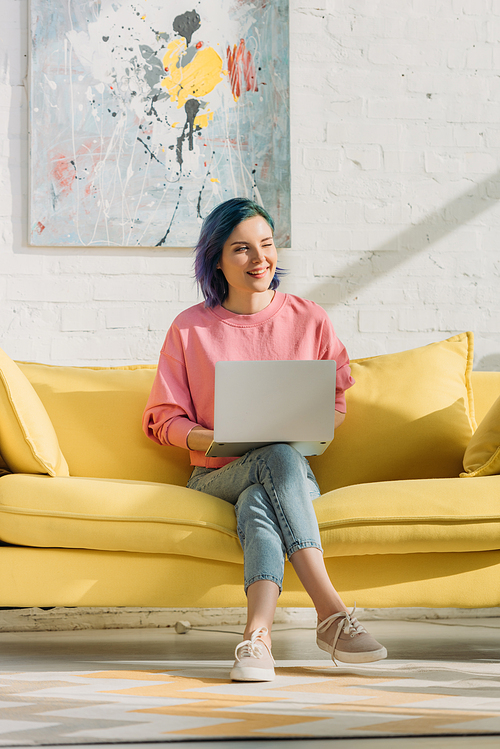 Freelancer with colorful hair and laptop smiling and sitting on sofa with crossed legs in living room