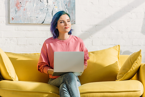 Freelancer with colorful hair and laptop looking away and sitting on yellow sofa with crossed legs in living room