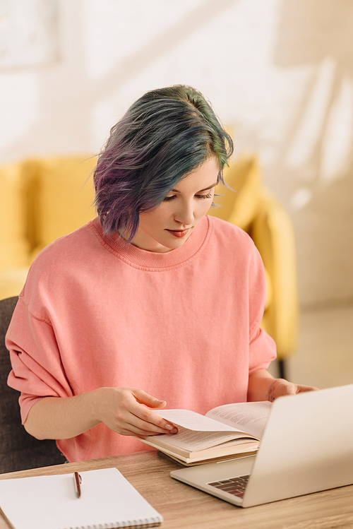 Freelancer with colorful hair reading book near laptop and copybook at table in living room