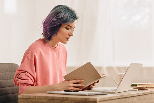 Freelancer with colorful hair reading book near laptop at table in living room