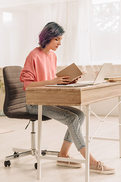 Freelancer with colorful hair reading book at table with laptop in living room