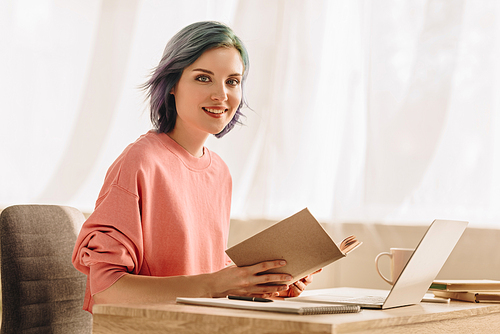 Freelancer with colorful hair and book  and smiling at table with laptop in living room