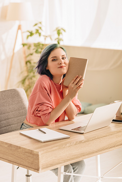 Freelancer with colorful hair holding book,  and smiling near laptop and notebook at table in living room