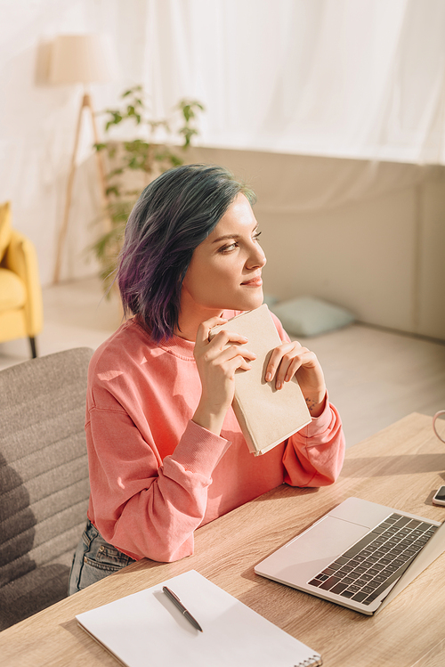 High angle view of freelancer with colorful hair holding book, looking away and smiling near laptop and notebook at table