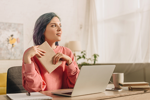 Freelancer with colorful hair and book looking away and smiling at table with laptop, notebook, cup of tea and smartphone in living room