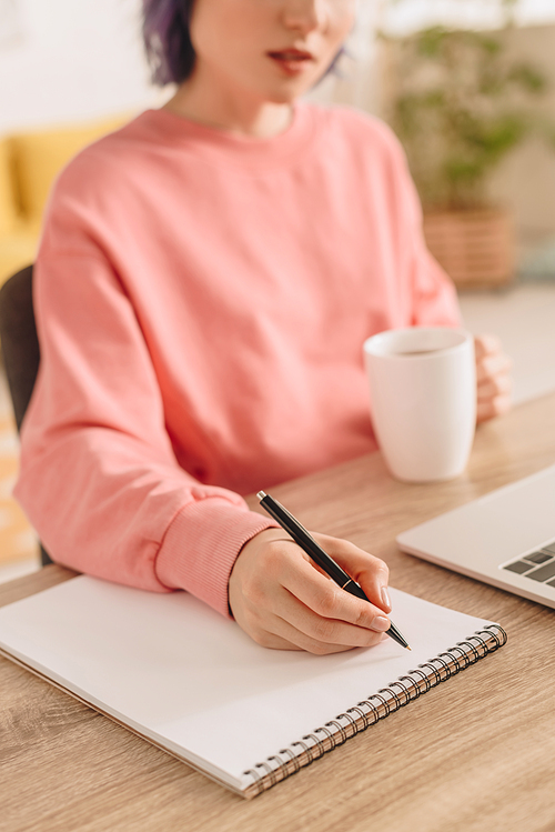 Cropped view of freelancer holding cup of tea and writing in notebook at table