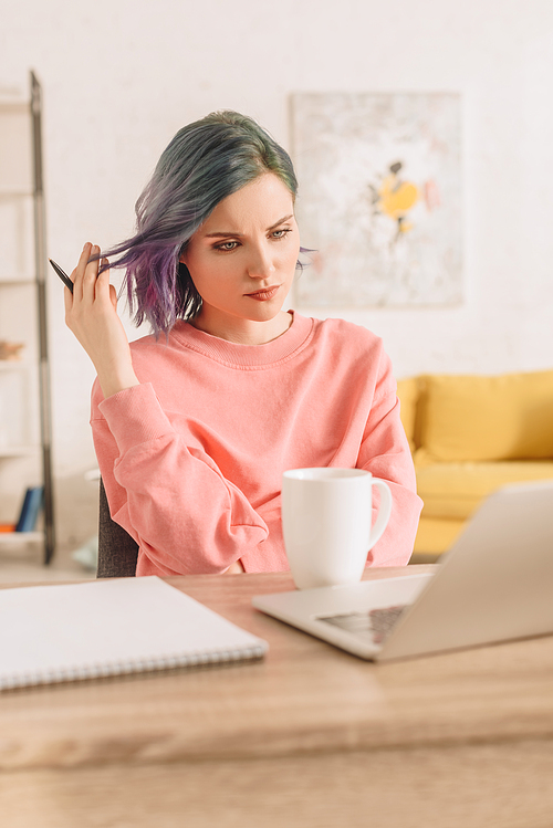 Selective focus of thoughtful freelancer with colorful hair holding pen at table with laptop, cup of tea and notebook in living room