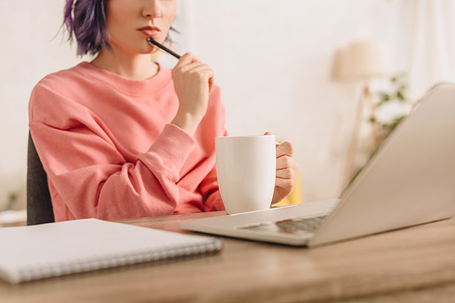 Cropped view of freelancer with colorful hair holding cup of tea and pen at table with notebook and laptop in living room