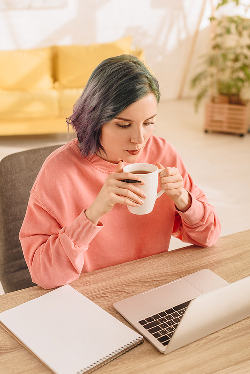 High angle view of freelancer with colorful hair holding cup of tea at table with laptop and notebook in living room