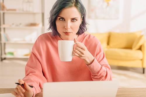 Selective focus of of freelancer with colorful hair  and holding cup of tea with pen at table in living room