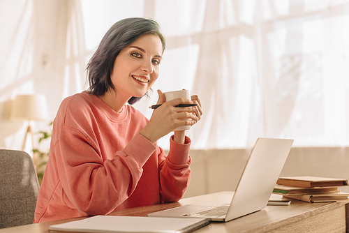 Freelancer with colorful hair, cup of tea and pen  and smiling at table with laptop in living room