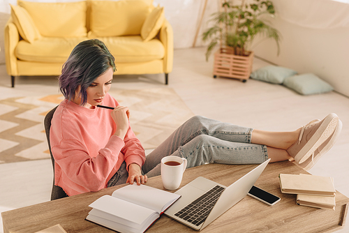 High angle view of freelancer with colorful hair and pen looking at notebook and putting legs on table