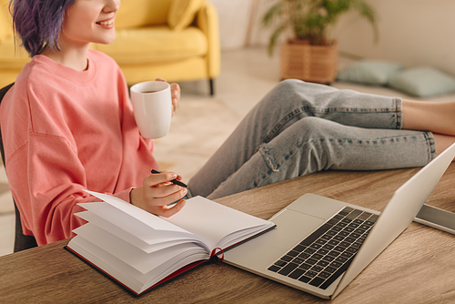 Cropped view of freelancer with colorful hair, cup of tea and pen putting legs on table with laptop and notebook in living room