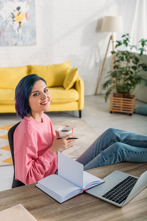 High angle view of freelancer with colorful hair, cup of tea and pen smiling and putting legs on table in living room