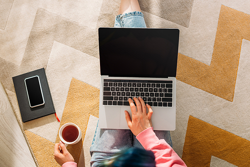 Cropped view of freelancer with cup of tea working with laptop near notebook and smartphone on floor in living room