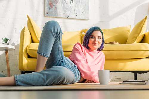 Selective focus of woman with colorful hair smiling,  and lying on floor near sofa with cup of tea in living room