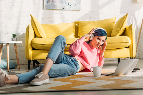 Freelancer with colorful hair and headphones looking at laptop and lying near sofa and cup of tea in living room