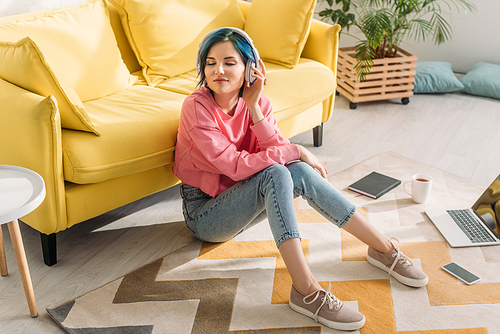 High angle view of freelancer with colorful hair, closed eyes and headphones smiling near sofa on floor